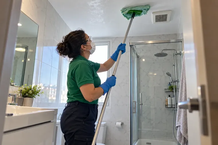 Gözlük ve maske takarak uzun saplı paspasla banyo tavanındaki küfü silmek